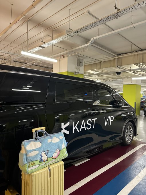 A black van labeled "KAST VIP" is parked in a garage, with a child-like bag featuring cartoon characters resting on a yellow suitcase.