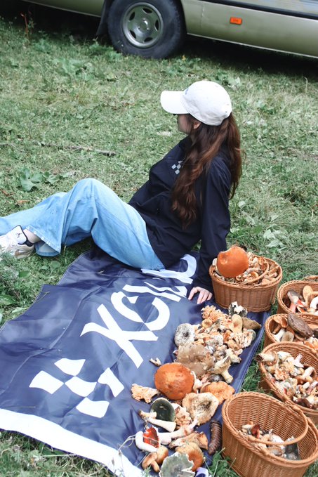A woman in a cap and dark jacket relaxes on a dark blue blanket spread on grass, surrounded by baskets filled with various mushrooms.
