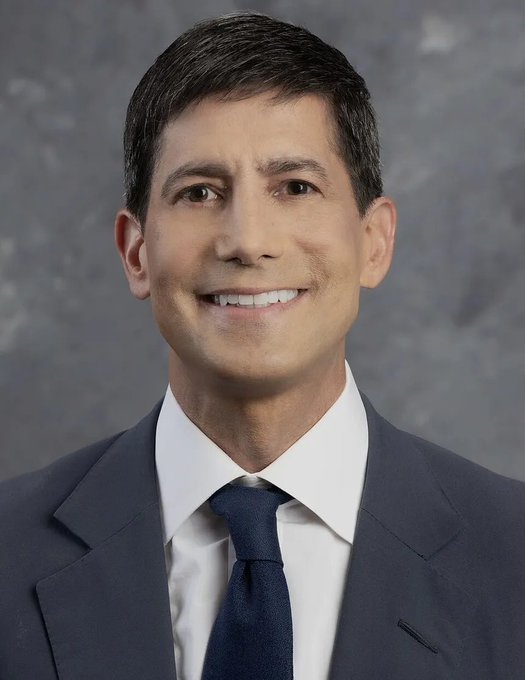 A professional headshot of a smiling man with dark hair wearing a navy suit, white shirt, and dark blue tie against a gray background.