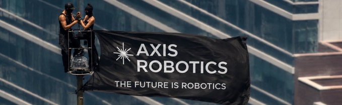 Two masked individuals on a raised platform unfurling a large black banner that reads 'AXIS ROBOTICS' and 'THE FUTURE IS ROBOTICS' against a backdrop of modern glass skyscrapers.