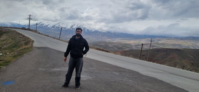 A man wearing a black jacket, gray pants, sunglasses, and a cap stands on a quiet mountain road with a sweeping view of snow-capped peaks and rolling hills behind him, perfectly capturing the serene start of a new week.