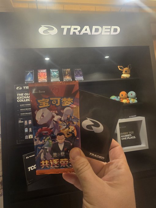 A hand holding a Chinese Pokémon trading card pack featuring Charizard alongside a black "TRADED" card sleeve, with a TV screen displaying "THE GLI EXCHANGE COLLECTION" and Pokémon plushies in the background.