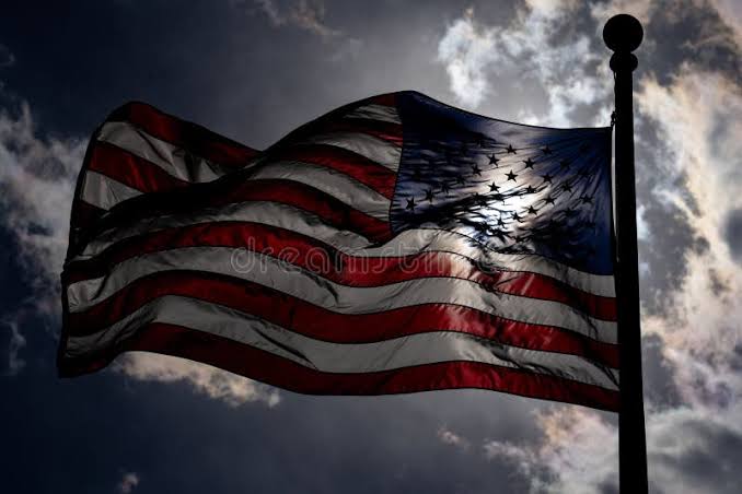 An American flag waves against a dramatic cloudy sky, backlit to create a striking silhouette effect.