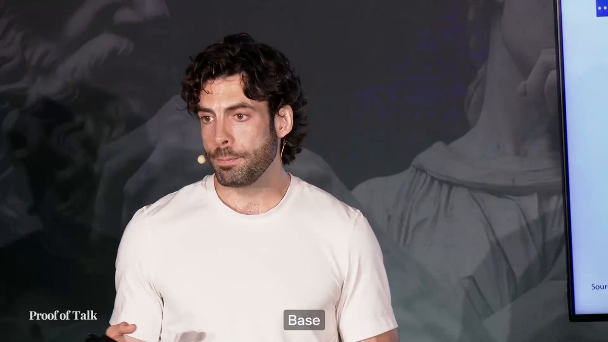 The image shows a man wearing a white t-shirt with a name tag that says "Base" on his chest. He appears to be giving a presentation or speech, as indicated by him standing behind a podium. The background of the image is black, which contrasts with the white t-shirt and makes it stand out more. There are also other people in the background who might be attending the same event.
