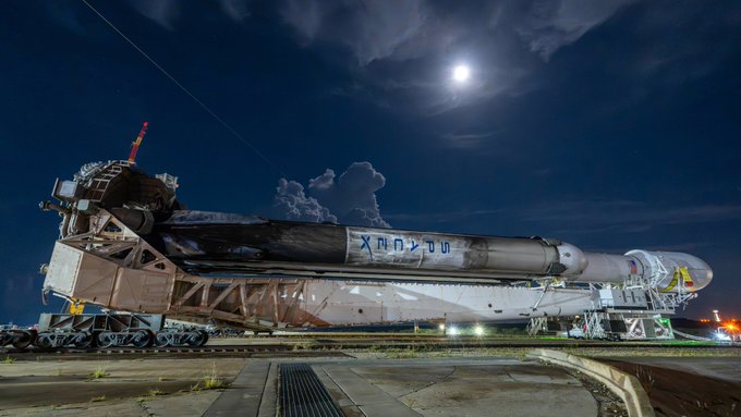 A SpaceX Falcon 9 rocket booster is transported horizontally on a raised platform at night, with the word "XODU" visible in graffiti on its side.