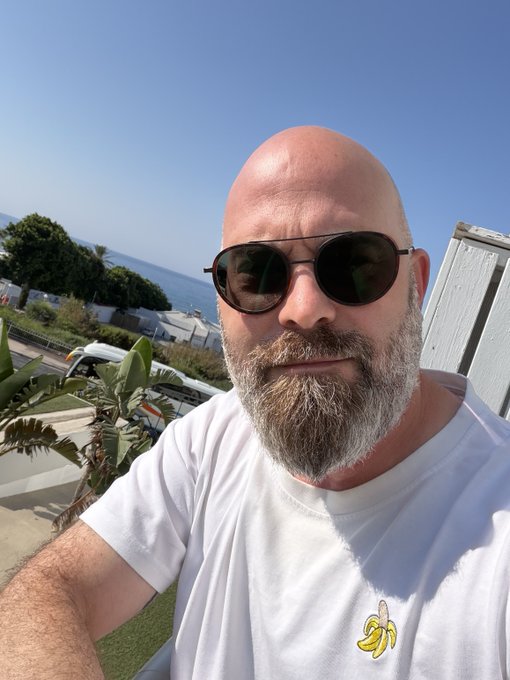 A selfie of a bald man with a gray beard wearing aviator sunglasses and a white t-shirt with a banana logo, taken outdoors with a coastal view in the background.