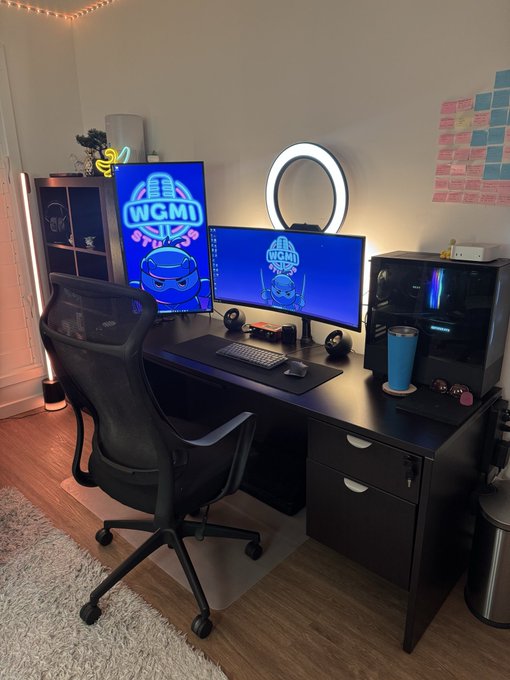 A dimly lit gaming setup featuring a black desk with two monitors and a PC displaying 'WGMI' logos with a cartoon frog, accompanied by a gaming chair, ring light, and sticky notes on the wall.