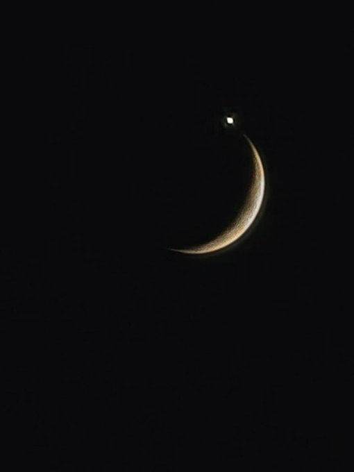 A photograph of a crescent moon in the dark night sky with a bright star or planet nearby.