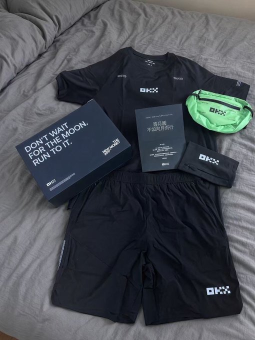 An athletic apparel set is laid out on a bed, featuring a black t-shirt, shorts, neon green belt bag, and wristband, all bearing the 'OKX' logo, alongside a box reading 'DON'T WAIT FOR THE MOON. RUN TO IT.'