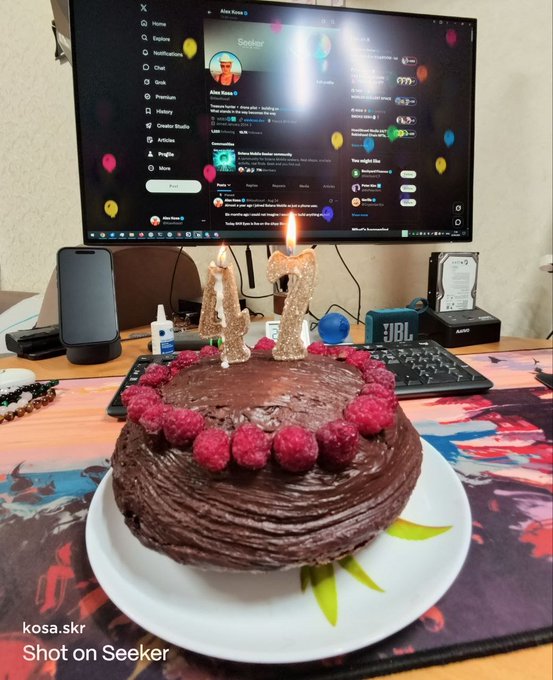 A chocolate cake decorated with raspberries and lit candles forming the number '42' sits on a desk in front of a computer monitor displaying the X (Twitter) profile for 'Aloe Xeno 0'.