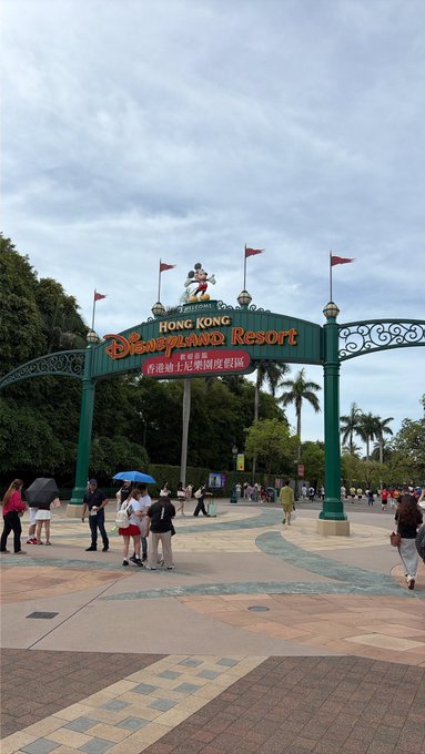 The entrance archway to Hong Kong Disneyland Resort with red flags, palm trees, and visitors walking around on a partly cloudy day.