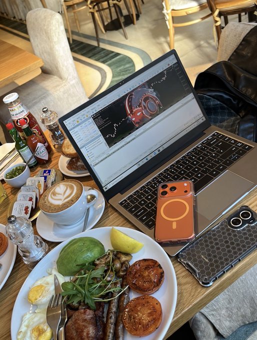 A café table holds an open laptop displaying a trading or charting application, a cup of latte, breakfast plates, phones, condiments, and other dining items.