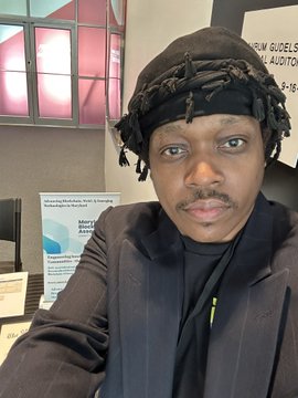 A selfie of a man wearing a black headwrap and black jacket, with a partially visible sign mentioning 'GÜEL' and 'AUDITOR' in the background.