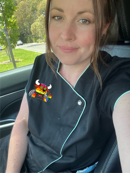 A woman sits in the driver's seat of a car wearing a black uniform with teal piping and an embroidered orange bull mascot logo, with greenery visible through the window.