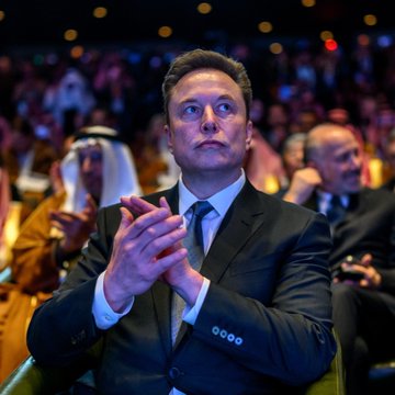 The image shows a man dressed in a suit sitting in an audience at a conference or event. The man appears to be listening intently to a speaker who is talking about the recent IPO of SpaceX, which has been putting pressure on Bitcoin as investors rotate their capital toward the offering.

In addition to the main subject, there are several other people present in the image, some sitting and others standing, indicating that it might be an event with multiple speakers or presentations taking place simultaneously. The presence of chairs suggests that this is a formal setting where attendees can sit comfortably while listening to the speaker.