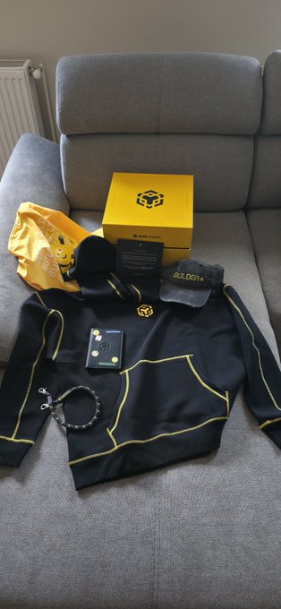 A collection of yellow and black themed merchandise items including a hoodie, a box, a cap, and accessories laid out on a gray sofa.