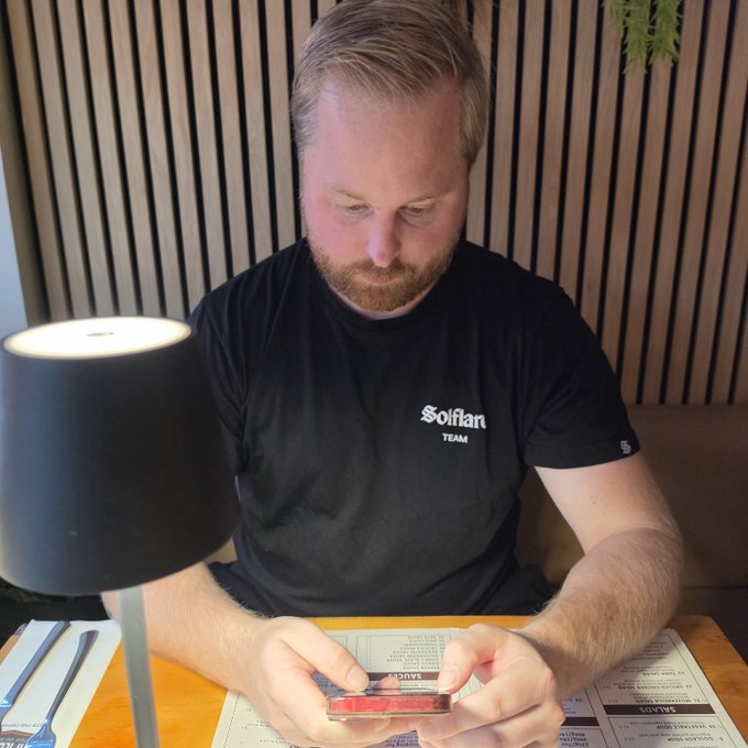 A bearded man wearing a "Solflare Team" t-shirt looks down at his phone while sitting at a restaurant table with menus in front of him, seemingly checking crypto markets instead of engaging with his date. The image humorously illustrates the post's complaint, showing him focused on Solana-related matters rather than his dining companion.