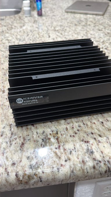 An IceRiver KAS Ultra ASIC miner sits on a granite countertop, ready to start mining Kaspa ($KAS) this weekend. The black heat-sinked device, labeled "IceRiver KS0 Ultra," represents the hardware for the user's first foray into KAS mining.