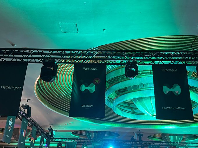 A vibrant event space bathed in teal lighting features multiple "Hyperliquid" banners hanging from metal trusses, including ones marked "VIETNAM" and "UNITED KINGDOM," confirming the international Hyperliquid gathering depicted in the post.
