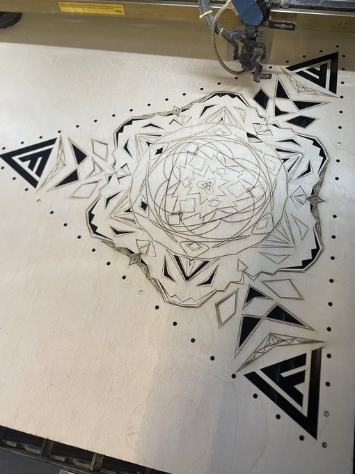 A laser cutter is engraving an intricate geometric mandala design onto a large wooden board, with a "LASER CAUTION" warning label visible on the machine. The intricate cut pattern—featuring triangles, diamonds, and a central rosette—is being prepared for display at WildoriaFest.