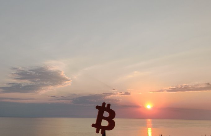 A sunset over water with a black Bitcoin (₿) symbol mounted as a silhouette in the foreground, featuring orange and pink sky colors.