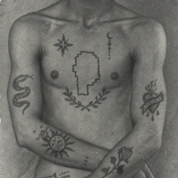 A black-and-white close-up showing a person's upper body adorned with various symbolic tattoos, including a snake, Sacred Heart, sun, star, and a pixelated face on the chest.