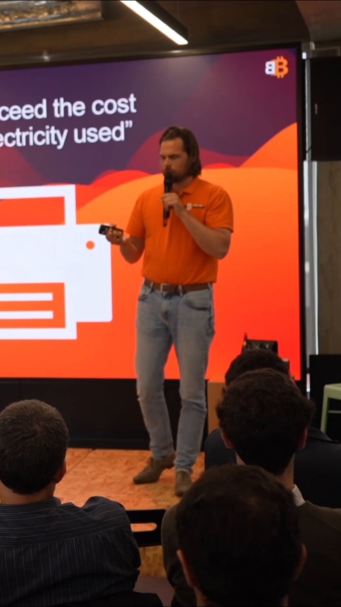 The image shows a man standing behind a podium, giving a speech or presentation to an audience. The room appears to be a conference hall with a large screen displaying information related to electricity usage. The speaker's attire consists of an orange shirt and blue jeans. The text on the screen reads "What is going to happen when they print a shit ton of money? Coming soon in war mongering EU." This suggests that the man might be discussing the potential consequences or implications of printing large amounts of money, possibly related to energy usage or other economic matters.