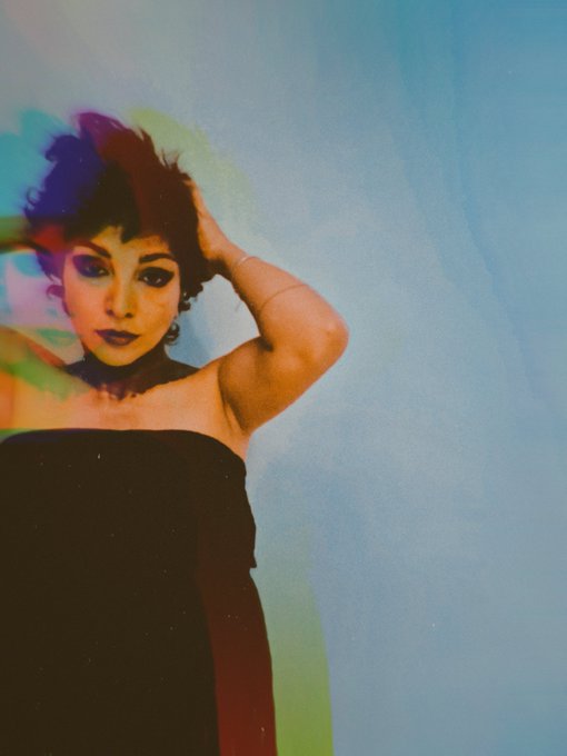 A stylized photo of a woman with dark hair and bold makeup wearing a black strapless dress, posed against a blue background with a rainbow color-leak effect.