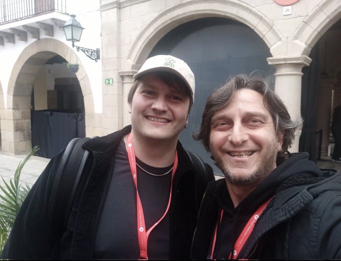 Two smiling men wearing red lanyards and casual jackets posing together for a photo in front of a stone arched building entrance, with one wearing a beige cap.