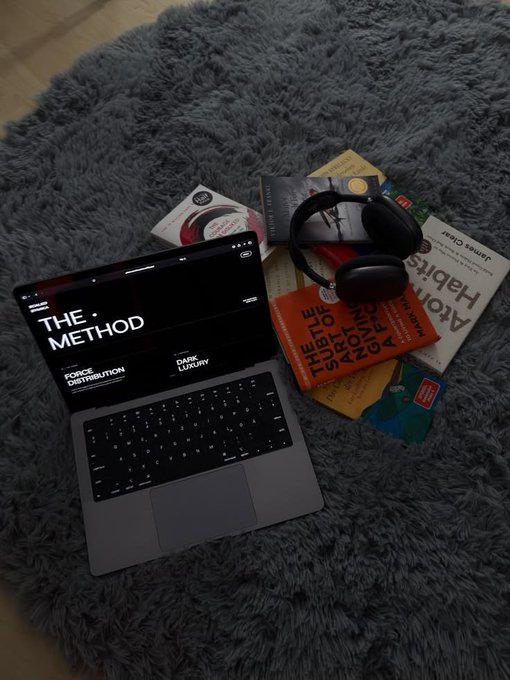An open laptop displaying 'THE METHOD' sits on a shaggy gray rug next to a stack of books including 'Atomic Habits' and 'A Philosophy of Software Design', topped with a pair of black headphones.