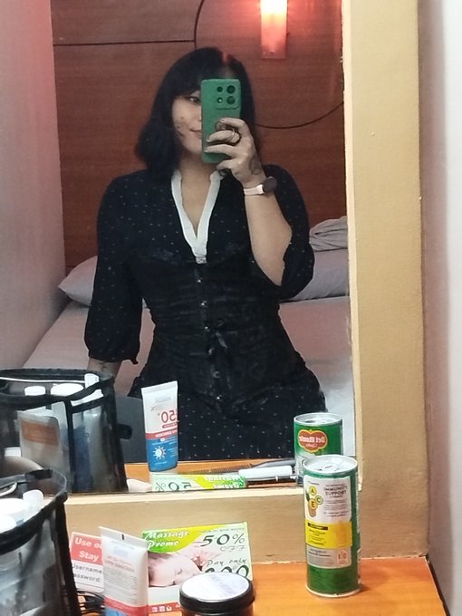 A mirror selfie of a woman in a black and white polka dot dress holding a green smartphone in a hotel bathroom, with various toiletries (including a 50% off sticker) visible on the counter.
