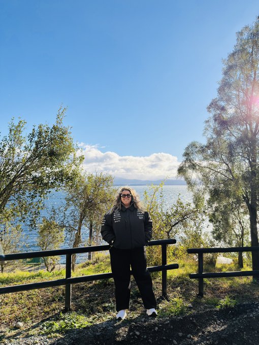 A blonde woman wearing sunglasses and a dark jacket poses for a photo in front of a scenic coastal lake and forested mountains under bright sunlight.
