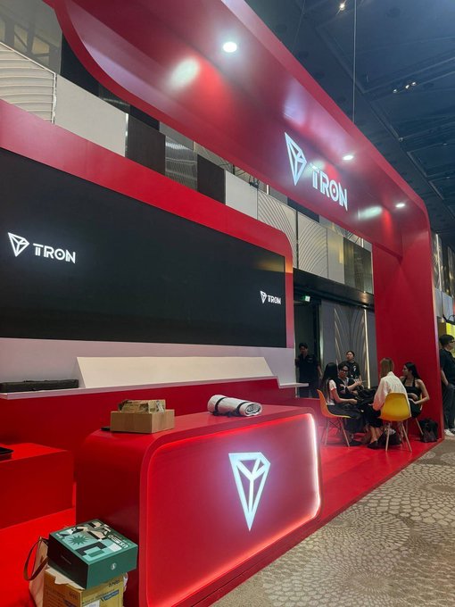 A red-themed TRON blockchain booth or event stand featuring the TRON logo on illuminated displays, with people seated at tables in the background.