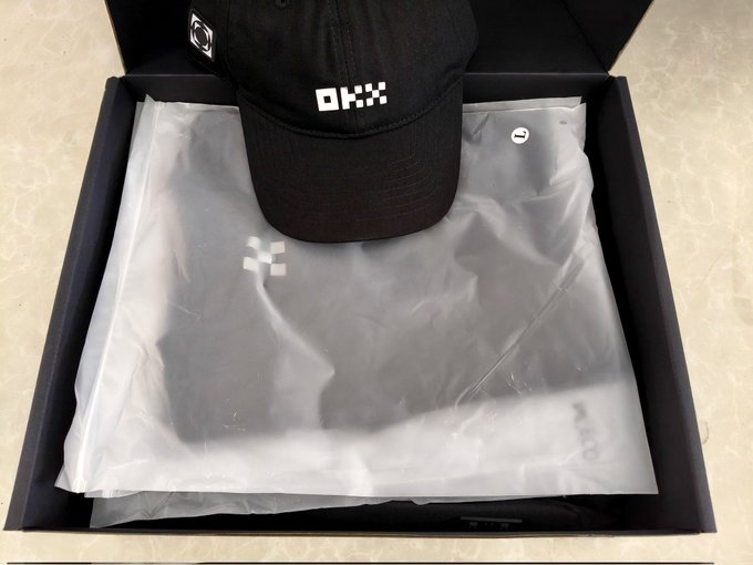 An OKX-branded black baseball cap displayed inside an open black gift box alongside tissue-wrapped merchandise.