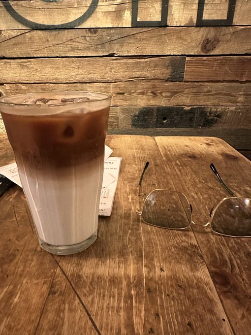 A glass of iced coffee sits on a wooden table next to a pair of reading glasses.