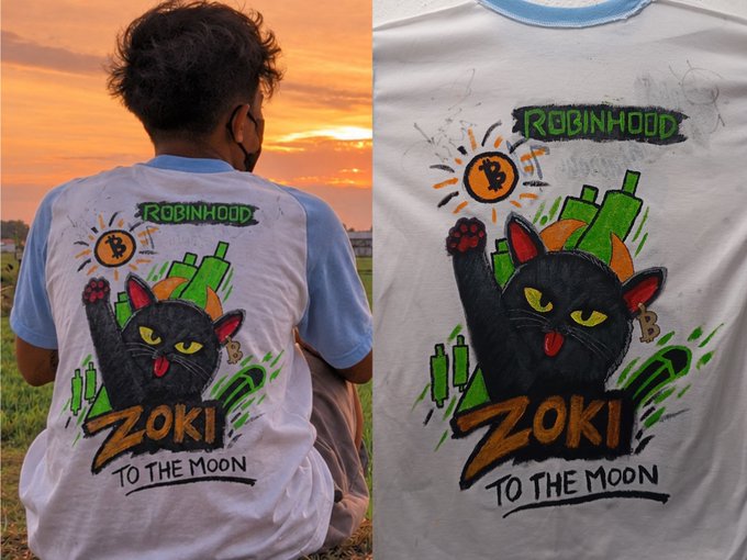 A child wearing a white t-shirt featuring a hand-drawn design of a black cat with 'ROBINHOOD' and 'ZOKI TO THE MOON' written on it sits in a field at sunset, shown next to a close-up of the artwork.