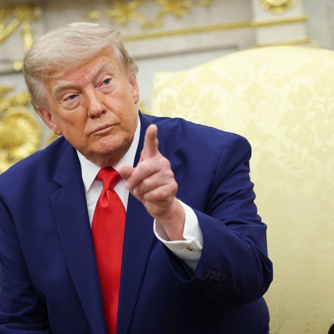 Donald Trump, dressed in a navy suit and red tie, gestures with a pointed finger while seated in an ornate room. The image relates to the article discussing his stock account activity involving Strategy (formerly MicroStrategy) and Tesla purchases.