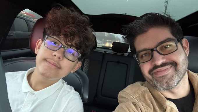 A selfie taken inside a car showing two people, one with curly brown hair wearing glasses and a white shirt, and another with a beard wearing glasses and a tan jacket.