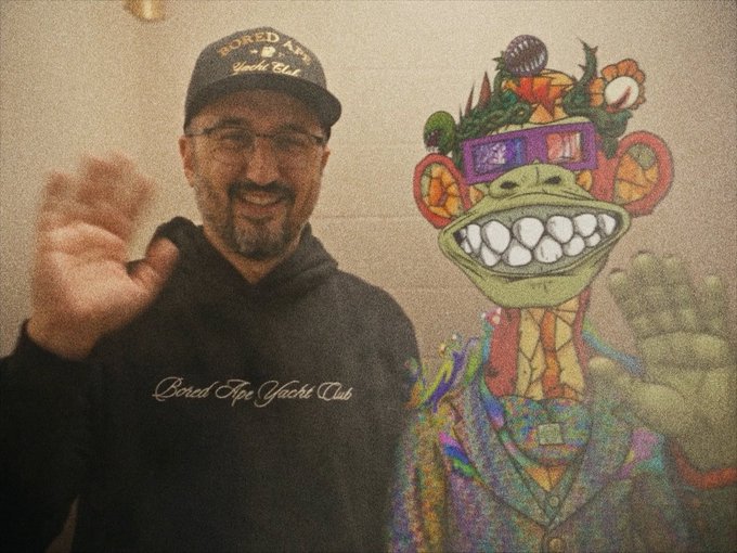A man wearing a 'Bored Ape Yacht Club' hoodie and matching cap waves at the camera, posing next to a colorful, psychedelic illustrated ape character with a wide grin.