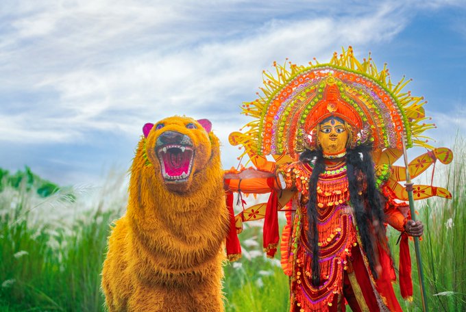 A person dressed in an elaborate, colorful traditional deity costume with a large ornate halo-like headpiece, standing next to a person wearing a furry orange and black bear costume in a grassy field.