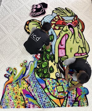 Two people wearing black hoodies (one labeled 'icd') are working on a large, colorful graffiti-style mural depicting a cartoonish green character on a concrete surface.