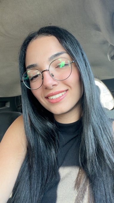 A selfie of a smiling young woman with long dark hair, round glasses, and a black top, sitting inside a car.