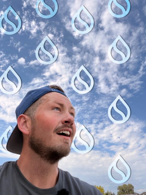 A young man wearing a blue backward baseball cap smiles and looks upward against a sky background filled with repeating Sui (SUI) cryptocurrency logos.