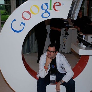 A man in a lab coat and glasses sits pensively inside a large Google "G" sculpture at what appears to be a tech conference. The Google branding and Bitcoin-focused caption about quantum computing resistance complement each other, suggesting the image represents a tech expert's perspective on cryptocurrency security within the broader tech industry context.