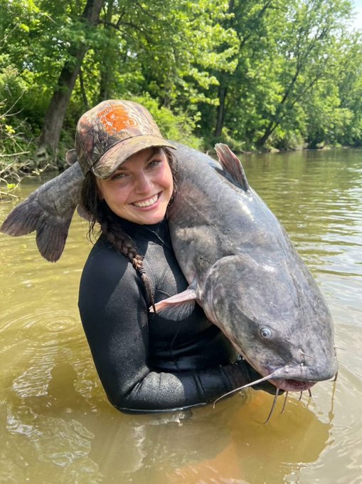 A smiling woman in a wetsuit and camouflage cap holds up a large catfish while standing waist-deep in a river.