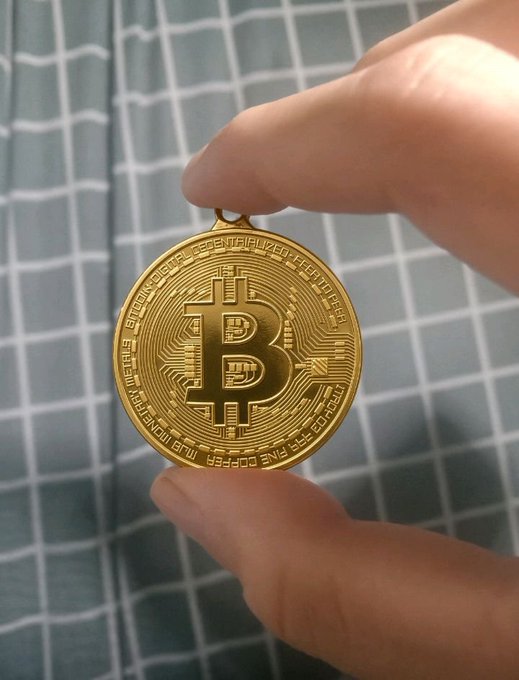 A close-up of a person's fingers holding a gold-colored Bitcoin commemorative coin with circuit board designs and the text 'BITCOIN DIGITAL DECENTRALIZED PEER TO PEER' and 'VIRES IN NUMERIS FINE COPPER'.