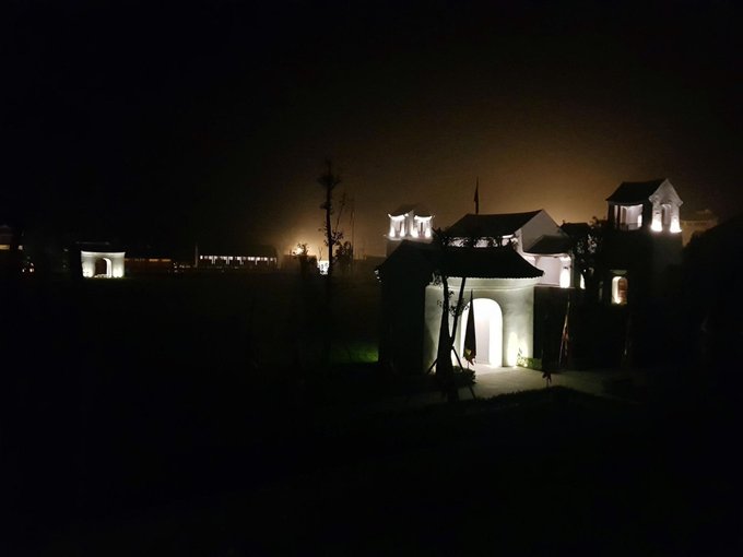 A nighttime photograph of a residential or compound area with several illuminated buildings featuring white walls and classical architectural elements, set against a dark sky with distant lights visible in the background.