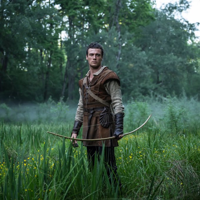 A man stands in a misty green meadow holding a wooden bow, dressed in medieval-style brown and beige clothing with leather arm guards.
