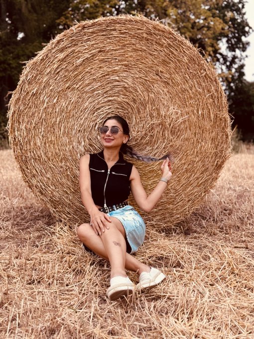 A woman in a black top, denim shorts, and sunglasses sits on dry grass in front of a large round hay bale, smiling with her braided hair held to the side.