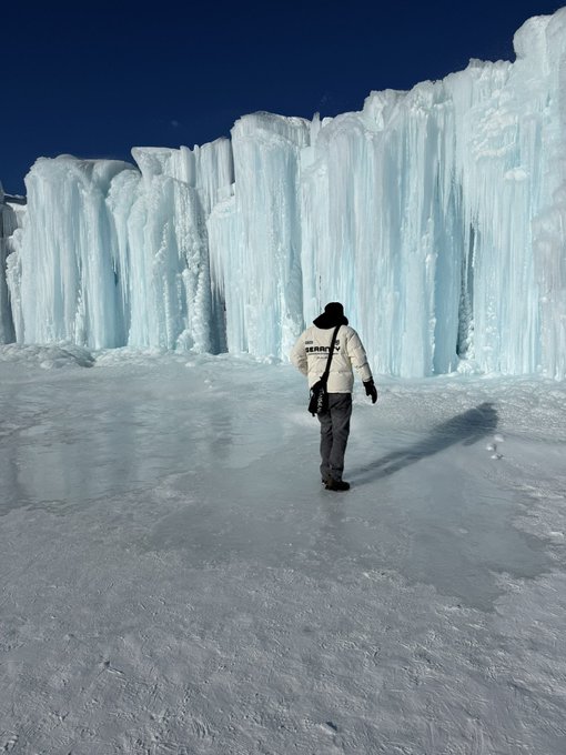 A person wearing a white Supreme jacket and black beanie walks across an icy expanse toward towering, translucent ice walls under a clear blue sky, with the brand name "Supreme" visible on the back of the jacket—showcasing the winter-ready style of the streetwear label.